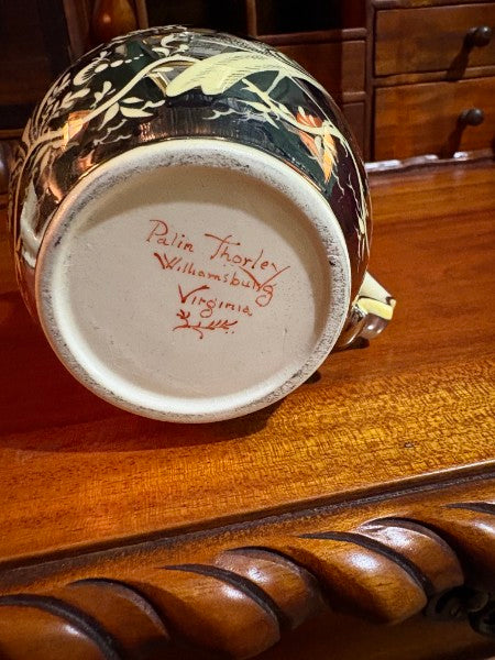 A close-up of the bottom of a decorative ceramic mug or cup, showing a cream-colored base with red hand-written signature text and a dark floral pattern around the rim, resting on a polished wooden surface.