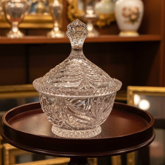 A clear cut-crystal glass lidded container with a decorative stopper, displayed on a round dark reddish-brown wooden side table against a gray corrugated metal background.