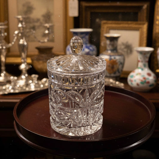 A clear cut-crystal glass jar with a matching lid, displayed on a dark brown wooden side table against a gray metal shutter background.