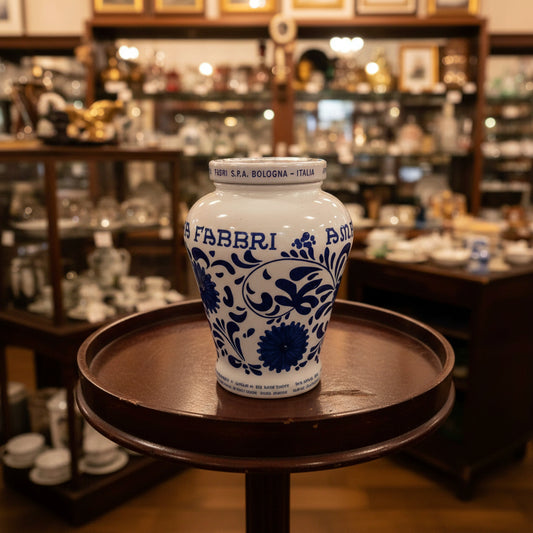A white ceramic Fabbri jar with blue floral patterns and the brand name 'FABBRI' on the front, sitting on a dark wooden stool in front of a white metal shutter.
