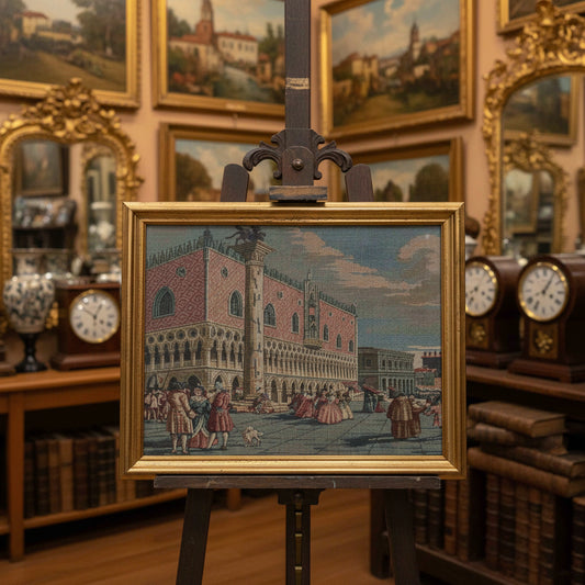 A hand holds a framed vintage-style painting depicting a grand architectural scene with figures in front of a large building. The frame is gold-toned, and the painting features muted pink, brown, blue, and beige tones.