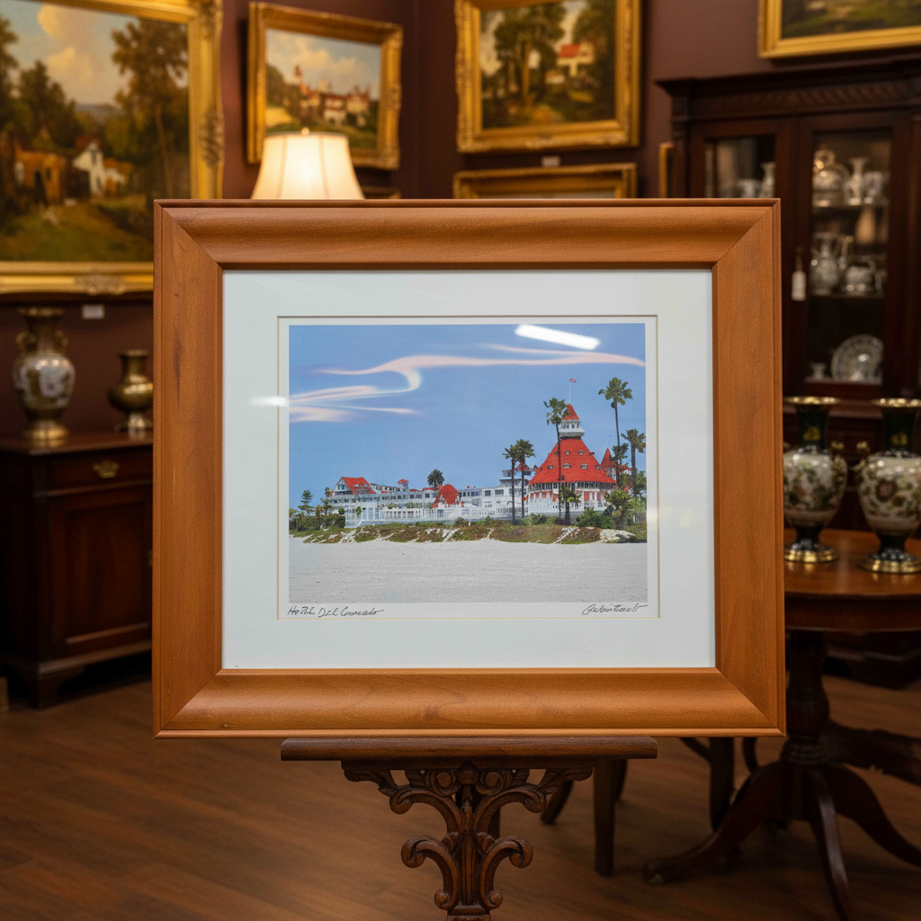 A framed art print depicting a beach scene with a lighthouse and white buildings under a blue sky, set in a wooden frame with a white mat, resting on a dark wooden table.