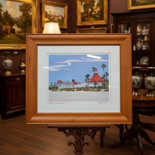 A framed art print depicting a beach scene with a lighthouse and white buildings under a blue sky, set in a wooden frame with a white mat, resting on a dark wooden table.