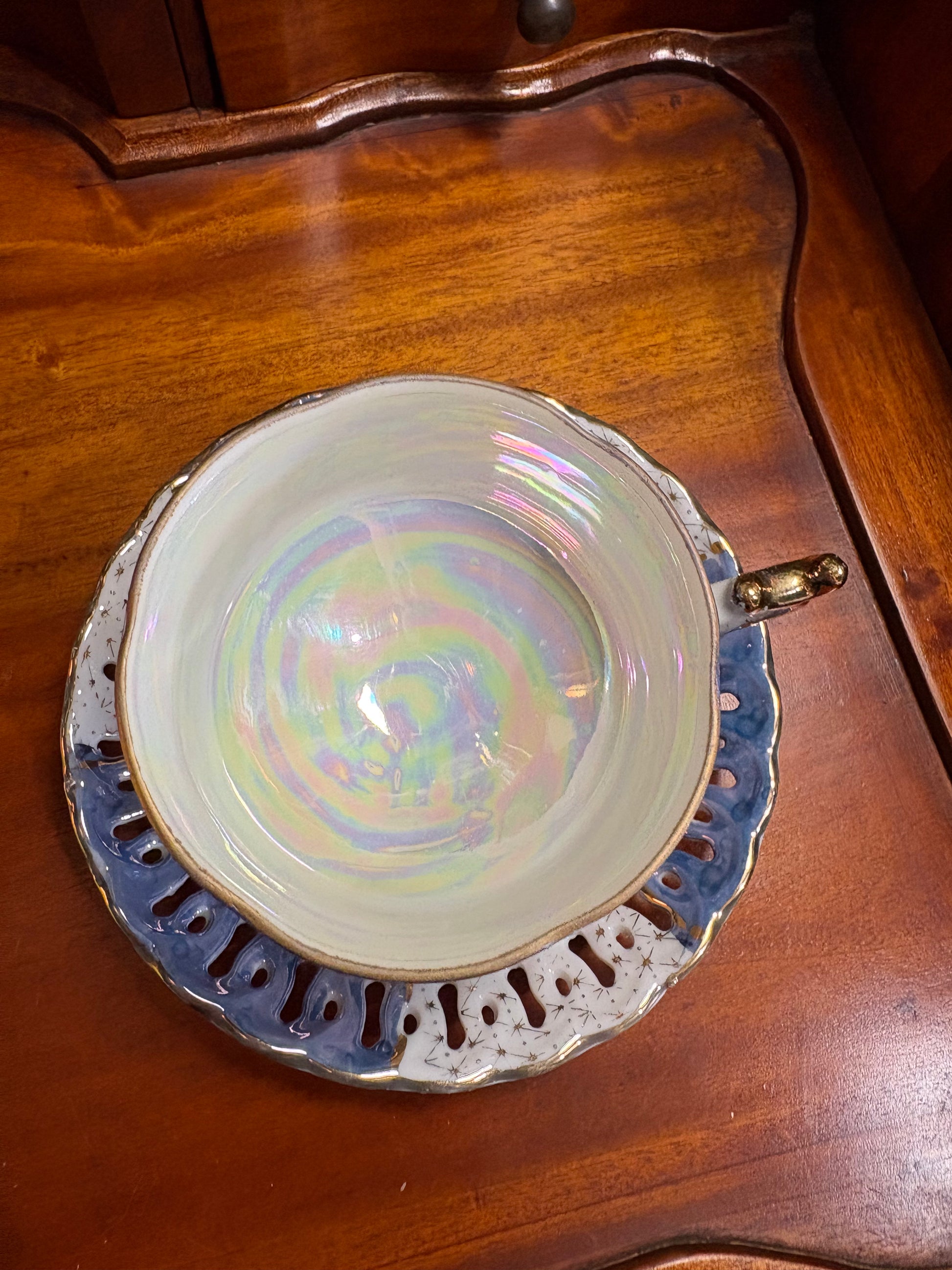An iridescent ceramic teacup and saucer set resting on a polished wooden table. The cup features a cream-colored interior with a swirling pastel rainbow sheen, a gold-toned rim, and a blue-gray scalloped saucer with cutout details.