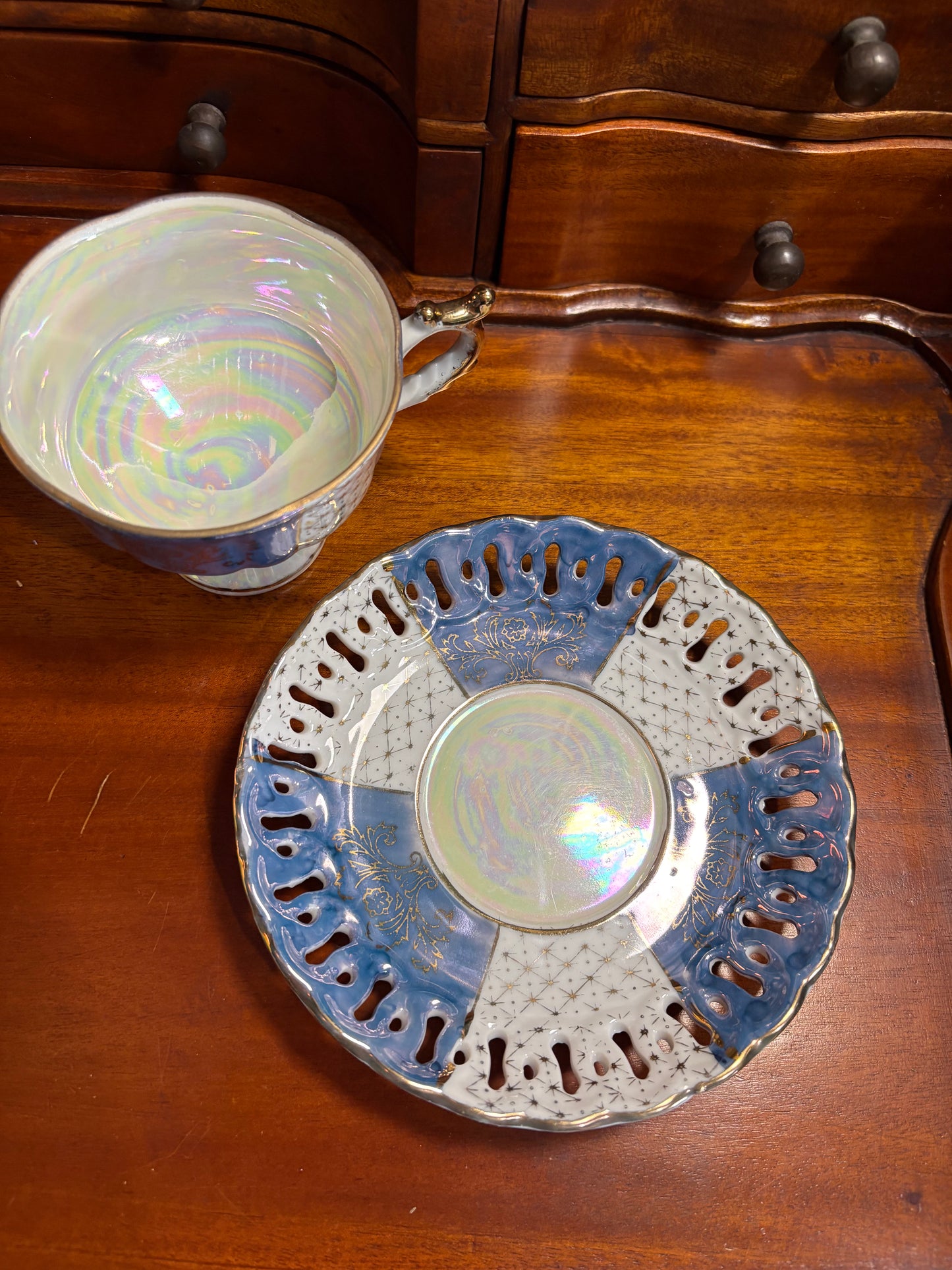 A vintage iridescent porcelain teacup and saucer set displayed on a polished wooden surface. The cup features a blue and white patterned exterior with a gold-toned rim, and the saucer has a scalloped blue edge with cutouts and a central iridescent white center.
