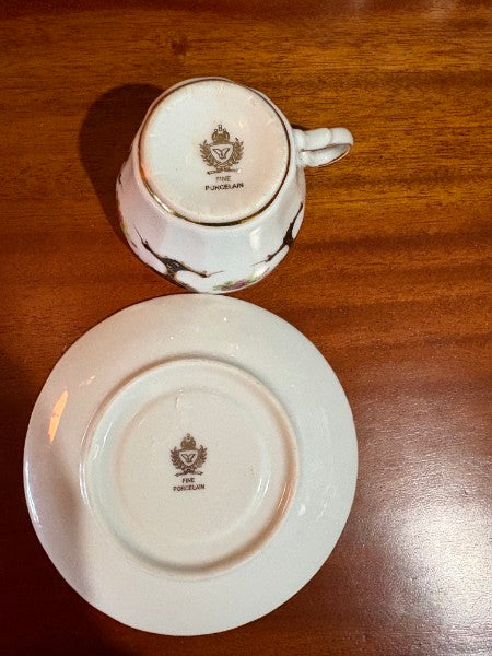 A white porcelain teacup and saucer set shown from the bottom, featuring a gold crest and 'THE PORCELAIN' text, placed on a wooden table.