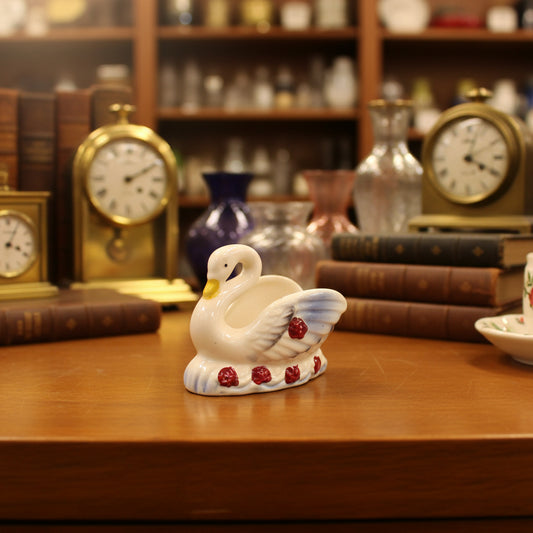 A small decorative swan figurine in pale pink with a yellow beak, white wings, and red heart accents on the tail, placed on a dark gray fabric background.