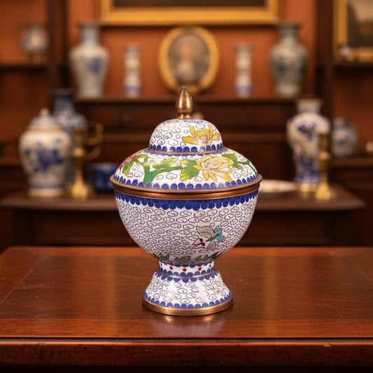 A small decorative lidded box with a pointed gold finial, decorated with white enamel, blue floral patterns, and yellow flowers, set against a dark green fabric backdrop.