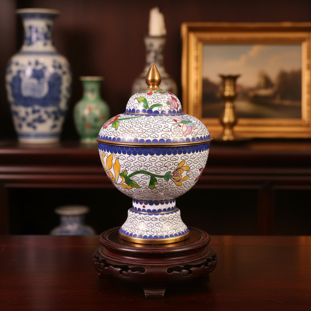 A decorative lidded ceramic jar or tumbler with intricate floral and scroll patterns, featuring a gold finial on top, set against a draped dark green fabric background.
