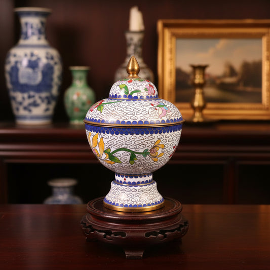 A decorative lidded ceramic jar or tumbler with intricate floral and scroll patterns, featuring a gold finial on top, set against a draped dark green fabric background.