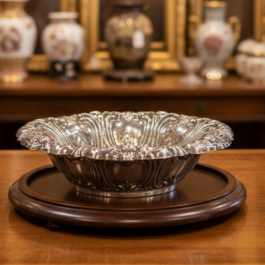 An ornate silver-colored metal bowl with a decorative embossed floral pattern, displayed on a draped green fabric background.