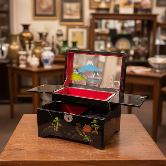 A black lacquered jewelry box with hand-painted green and orange floral motifs, shown open to reveal a bright red velvet-lined interior and a small mirror on the inside lid, set against a draped gray fabric backdrop.