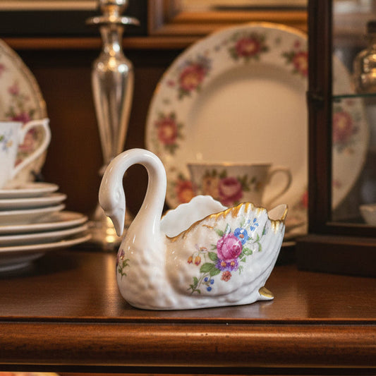 A small white swan-shaped ceramic pitcher with a floral pattern and gold trim, set against a dark green fabric backdrop.