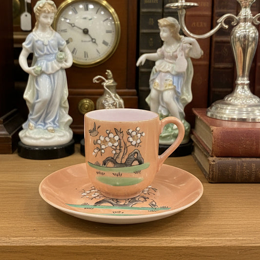 A small pink ceramic teacup and saucer set with a floral and landscape illustration, placed on a dark gray fabric backdrop.