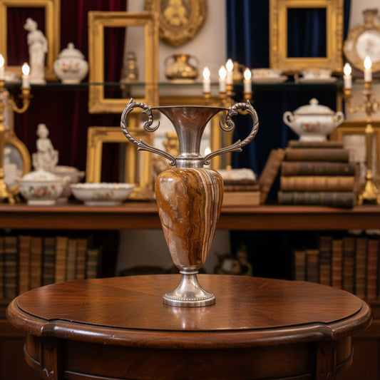 A decorative urn-style vase with a polished silver-toned metal body, double scrolled handles, and a marbled orange and cream stone or glass body, set against a draped dark gray fabric backdrop.