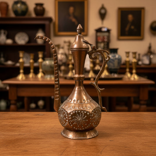 An ornate copper Arabic tea or coffee pitcher with a long curved spout and decorative handle, set against a dark green fabric backdrop.