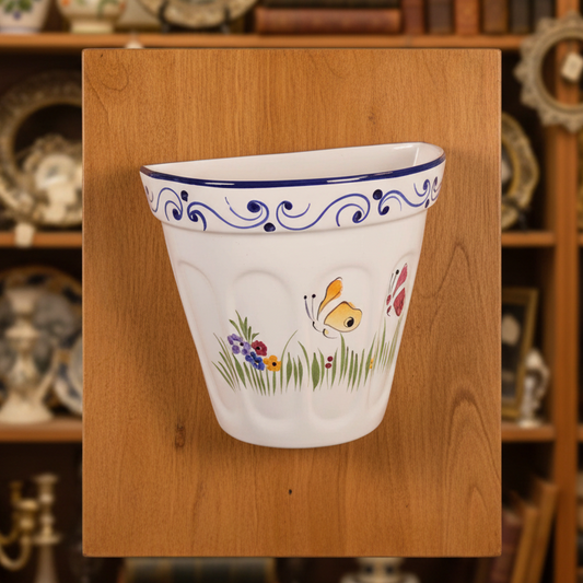 A white ceramic wall-mounted planter with blue scrollwork trim and a painted Easter-themed design featuring a butterfly, flowers, and a chick, set against a dark draped fabric background.