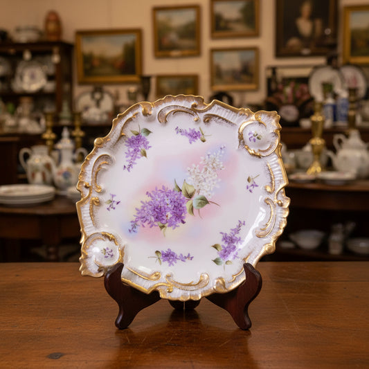 An ornate, scalloped porcelain plate with purple floral patterns and gold trim, displayed on a draped dark gray fabric background.